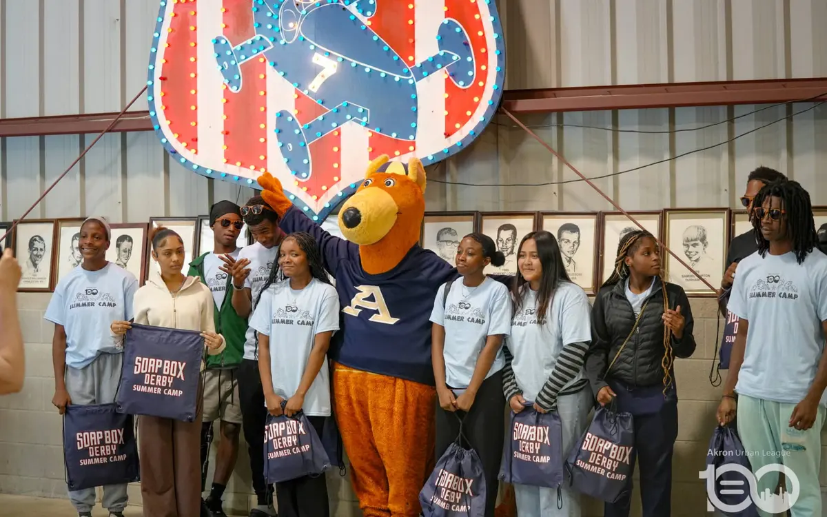 Youth participants and mascot smiling together holding tote bags at the Soap Box Derby summer camp
