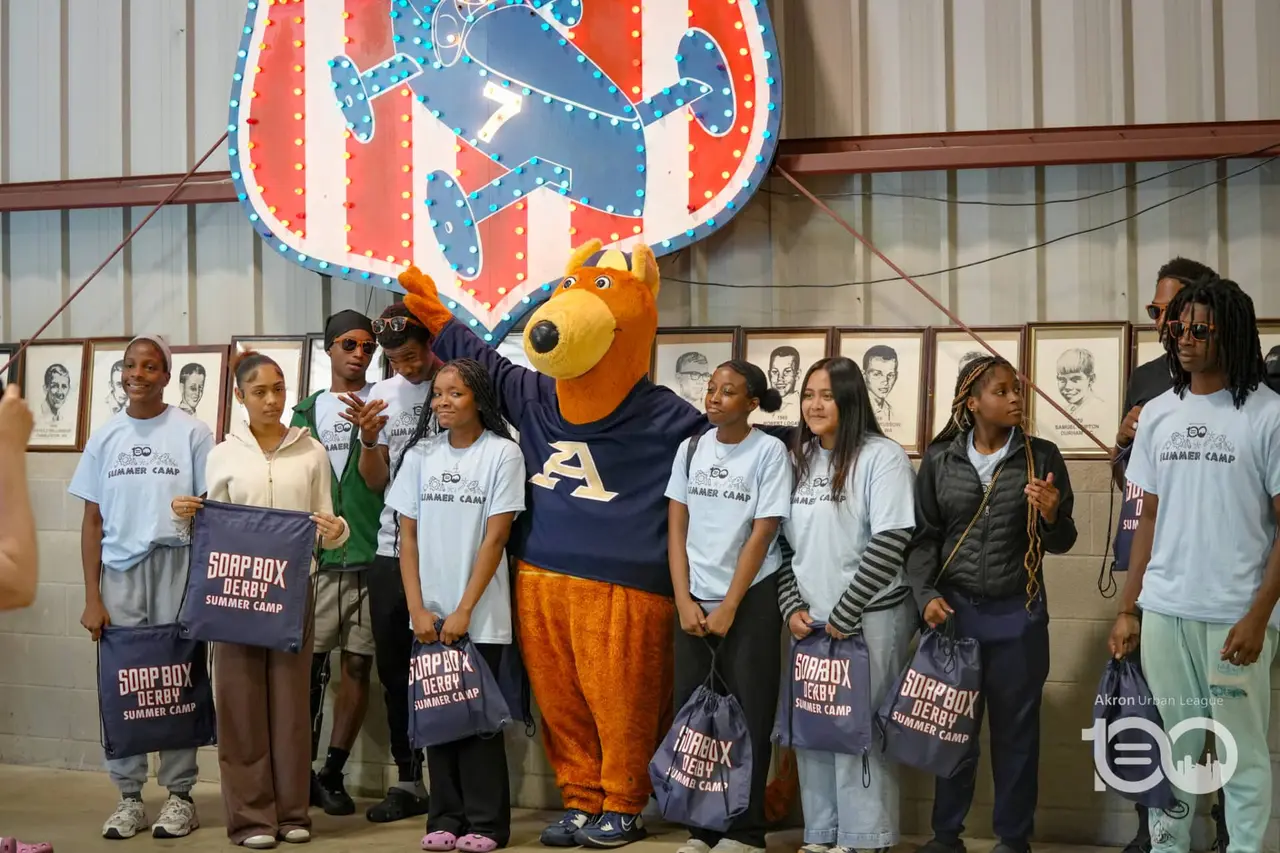 Youth participants and mascot smiling together holding tote bags at the Soap Box Derby summer camp