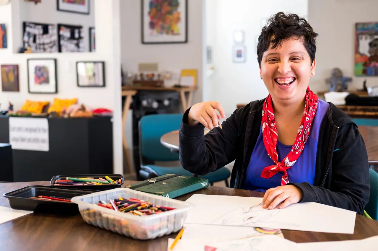 An artist smiling at a table with colored pencils at the Slingshot art studio