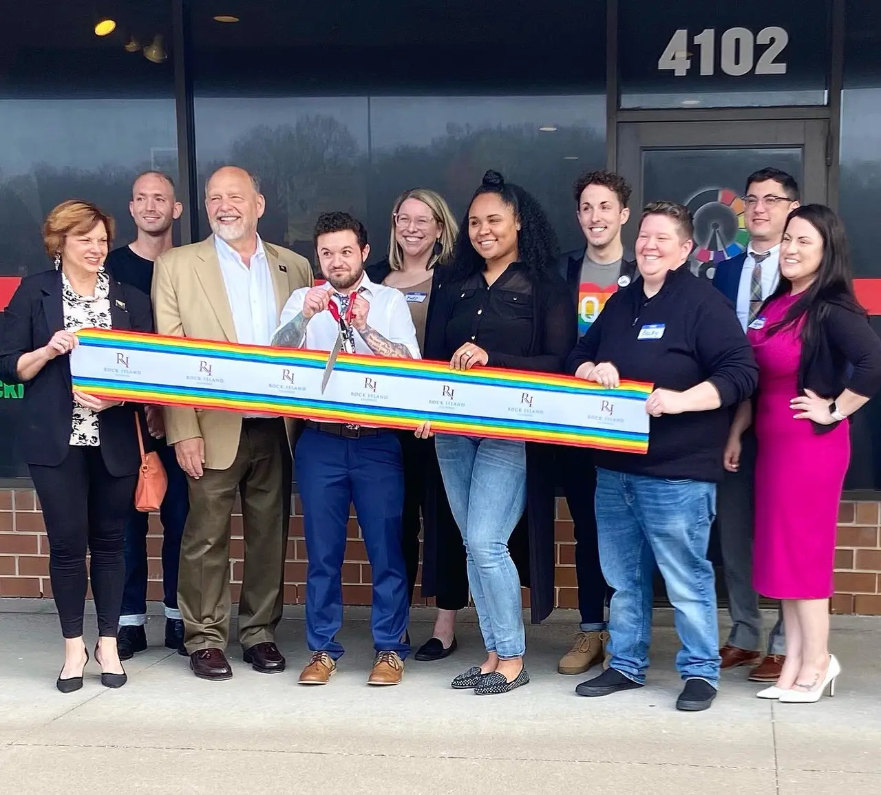 Staff, leadership, and local supporters holding a ceremonial rainbow ribbon outside the Clock, Inc. community center