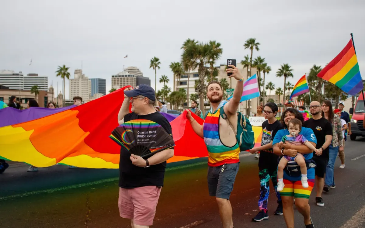 Marchers carrying a giant rainbow flag during an outdoor community Pride parade in Corpus Christi