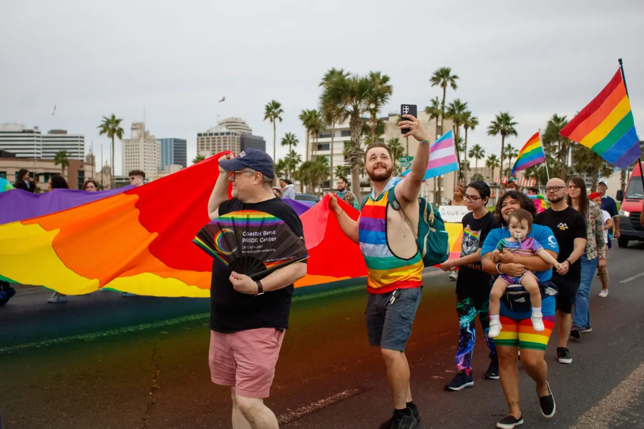 Marchers carrying a giant rainbow flag during an outdoor community Pride parade in Corpus Christi