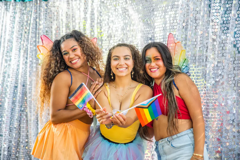 Three friends posing with small rainbow flags in front of a sparkling backdrop at Honolulu Pride