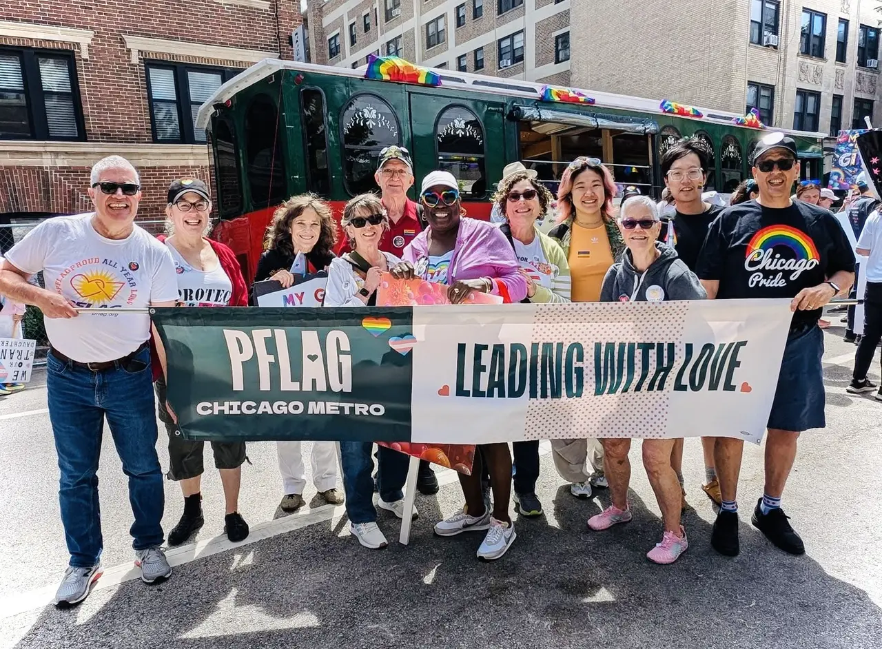 Ten PFLAG Chicago Metro members holding a banner at the Chicago Pride Parade with a PFLAG trolley behind them