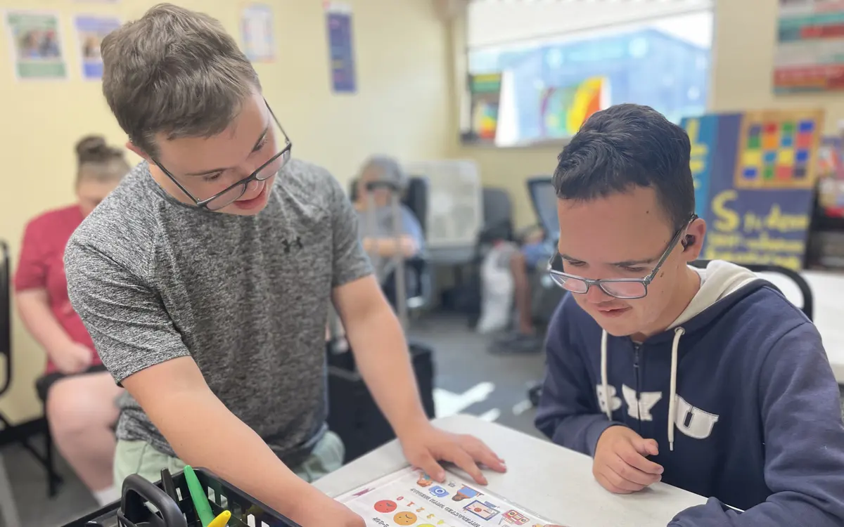 Two youth participants smiling and collaborating over an activity check-in sheet at a learning table