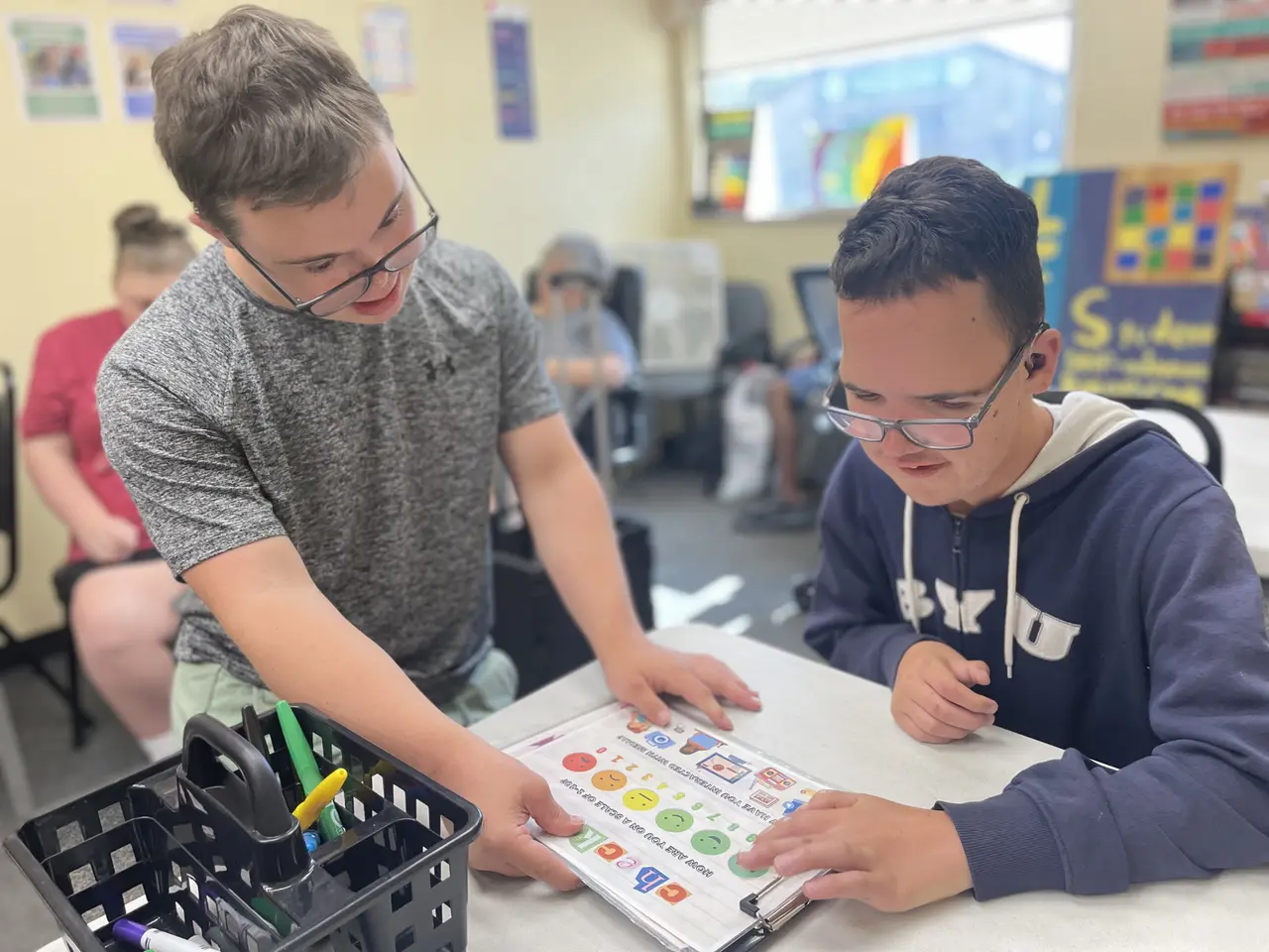 Two youth participants smiling and collaborating over an activity check-in sheet at a learning table