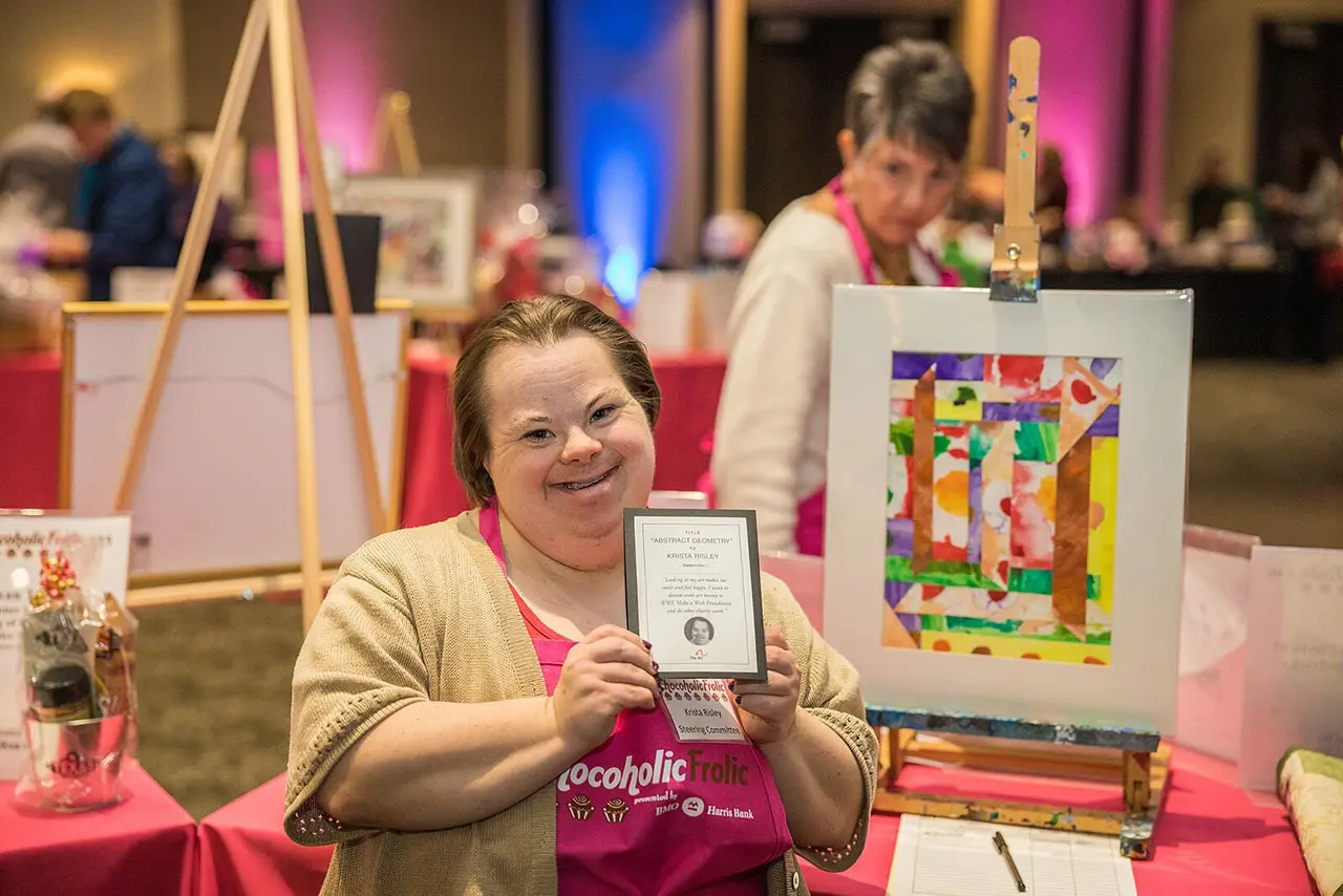An artist with Down syndrome smiling proudly while displaying her painting and award certificate