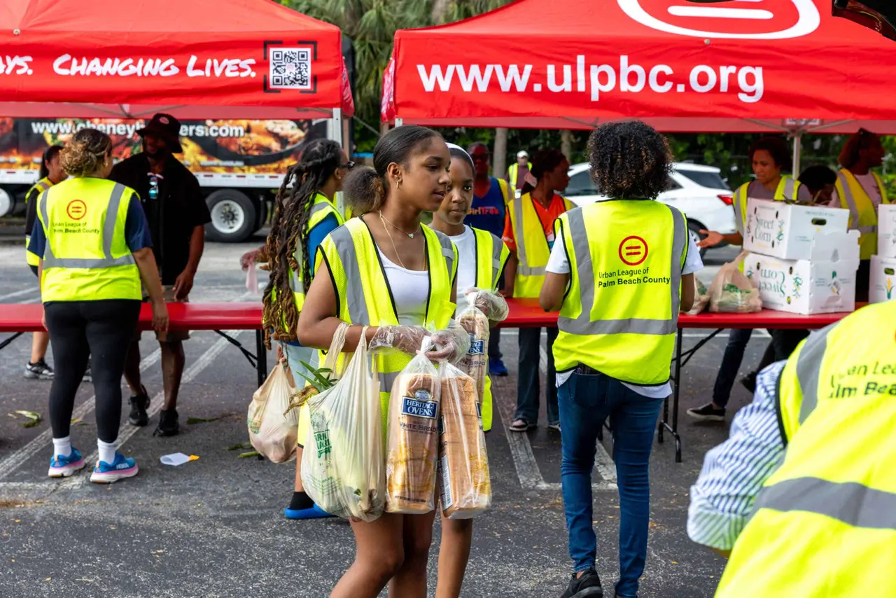Volunteers in safety vests carrying bags of food under Urban League tents at the 2026 Summer Food Drive