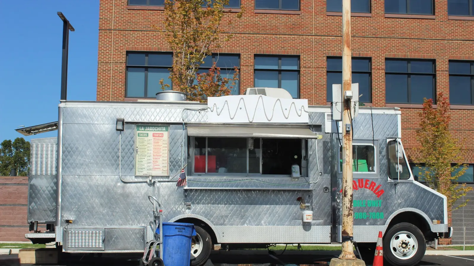 A silver taco truck with a hand-painted menu board, parked in front of a brick building