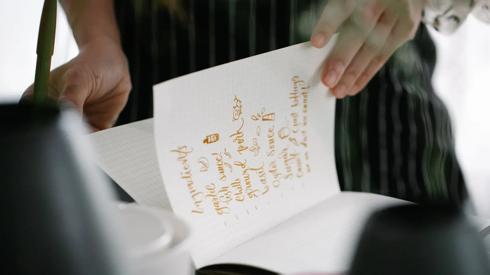 Hands holding open a notebook with an ingredient list handwritten in gold ink