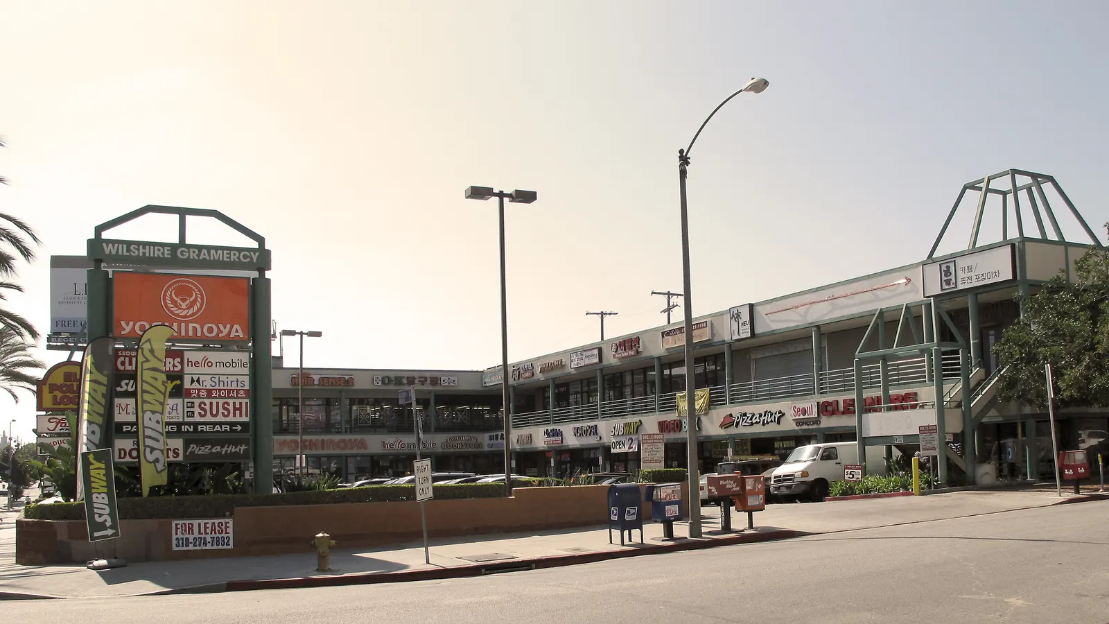 A two-story corner mini-mall on Wilshire Boulevard in Los Angeles's Koreatown, its pylon sign stacked with a fast food chain, a sushi bar, dry cleaners and Korean-language signs