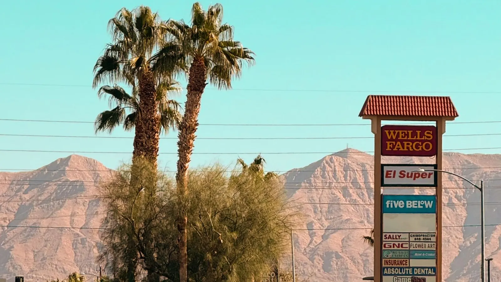 A tall shopping center pylon sign against desert mountains, with a bank, a Latino supermarket, a discount store and a column of smaller tenants including a dentist