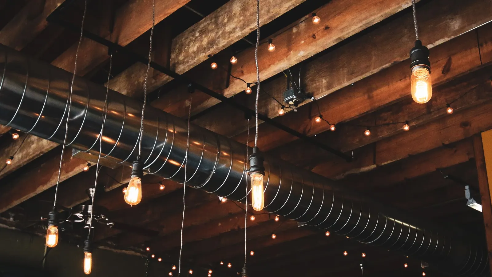 Exposed ductwork and wooden beams across a restaurant ceiling, strung with bare bulbs and hanging lights