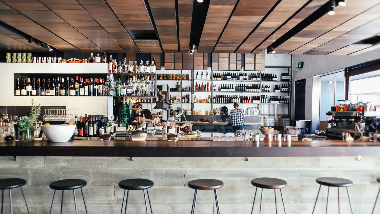 A long concrete bar with a row of backless metal stools, shelves of bottles and a hard, bright room behind it